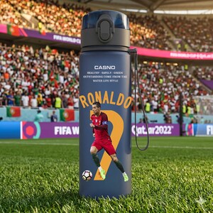 May include: A navy blue water bottle featuring a soccer player graphic and the name "Ronaldo." The bottle has a black cap and a strap. Text on the bottle reads "CASNO" and "HEALTHY - SAFELY - GREEN OUTSTANDING COME FROM THE WATER LIFE STYLE."