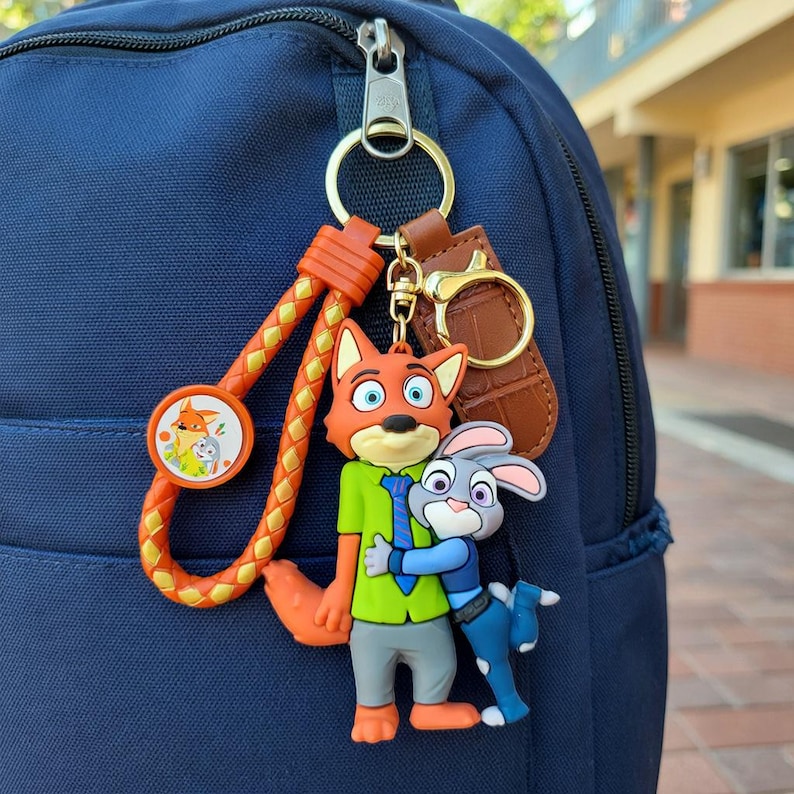 May include: A Zootopia-themed keychain featuring Nick Wilde and Judy Hopps characters. The keychain has an orange braided strap and a brown leather tag. The characters are in vibrant colors, attached to a blue backpack.