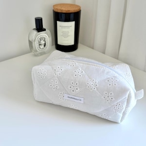 May include: A white quilted cosmetic bag featuring an eyelet floral pattern and a zip closure. A small white rectangular label with the black text Amy&Many.Co is sewn onto the front. The bag has a soft, padded texture and a rectangular shape.