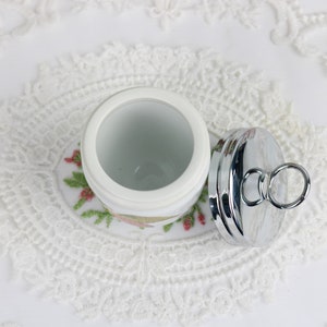 May include: A white ceramic jar with a floral design, paired with a shiny silver lid. The lid has a ring handle. The jar sits on a white lace doily, creating a vintage aesthetic.