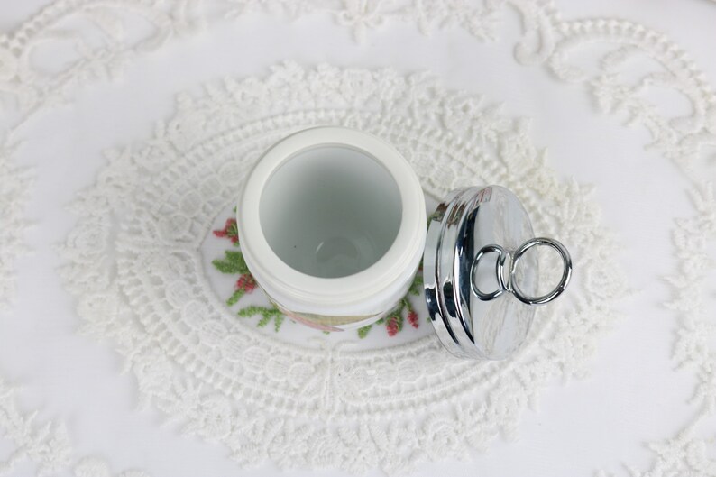 May include: A white ceramic jar with a floral design, paired with a shiny silver lid. The lid has a ring handle. The jar sits on a white lace doily, creating a vintage aesthetic.
