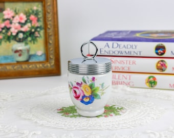 Puede incluir: Un huevo de cerámica blanca con un diseño floral y una tapa plateada con un asa de anillo. El huevo está sentado sobre un mantel de encaje blanco.