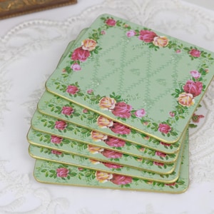Puede incluir: Una pila de cinco posavasos cuadrados con un fondo verde claro y un diseño floral. Los posavasos tienen un borde dorado y están decorados con rosas rosas y amarillas. El fondo tiene un sutil patrón de diamantes.
