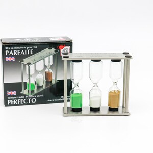 May include: A stainless steel tea timer with three hourglasses, each containing a different colored sand: green, white, and tan. The timer is in a rectangular frame, with a box in the background that reads "PARFAITE" and "PERFECTO".