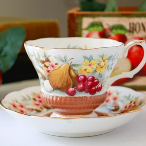 Puede incluir: Una taza de té y un platillo blancos con un patrón floral que presenta peras, cerezas y flores amarillas. La taza y el platillo tienen un borde dorado.