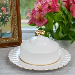 May include: White ceramic butter dish with a gold knob on the lid and a gold trim around the edge of the plate. The dish is sitting on a gray tablecloth.