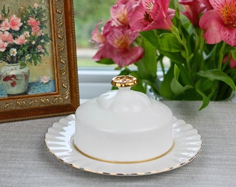 May include: White ceramic butter dish with a gold knob on the lid and a gold trim around the edge of the plate. The dish is sitting on a gray tablecloth.