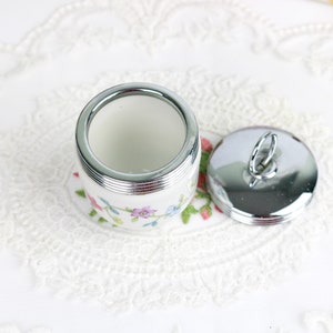K&ouml;nnte beinhalten: Eine wei&szlig;e Keramik-Eierbecher mit Blumenmuster und einem silbernen Deckel mit einem Schlaufe. Der Eierbecher steht auf einer wei&szlig;en Spitzenunterlage.