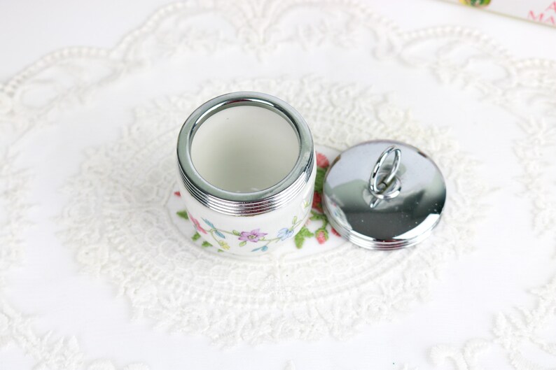 K&ouml;nnte beinhalten: Eine wei&szlig;e Keramik-Eierbecher mit Blumenmuster und einem silbernen Deckel mit einem Schlaufe. Der Eierbecher steht auf einer wei&szlig;en Spitzenunterlage.
