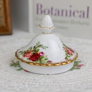 Op de afbeelding: Een witte porseleinen deksel met een gouden rand en een bloemenrozenontwerp. Het deksel heeft een kleine, afgeronde knop en is versierd met rode en roze rozen en groene bladeren. Een decoratief item.