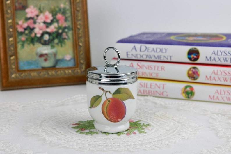 May include: White ceramic egg cup with a silver-tone lid and handle. The egg cup features a colorful illustration of a peach with green leaves. The egg cup is sitting on a white lace doily.
