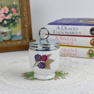 May include: A white ceramic egg cup with a silver lid and handle, decorated with a blackberry design. The egg cup is on a small, embroidered doily. Books and a framed picture are in the background.