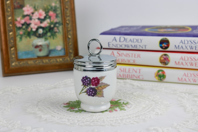 May include: A white ceramic egg cup with a silver lid and handle, decorated with a blackberry design. The egg cup is on a small, embroidered doily. Books and a framed picture are in the background.