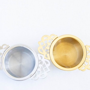 May include: Two tea infusers with intricate, decorative handles. One infuser is silver and the other is gold.