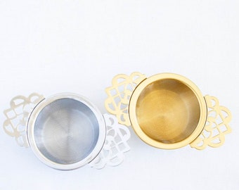 May include: Two tea infusers with intricate, decorative handles. One infuser is silver and the other is gold.