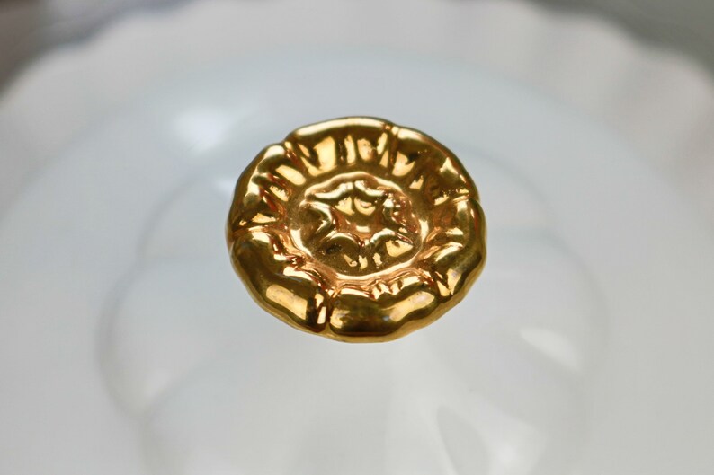 May include: A gold-colored, decorative knob with a floral design. The knob is round and has a raised center.