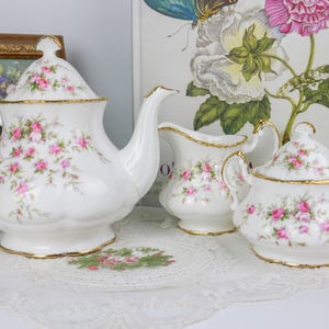 Peut inclure: Service à thé en porcelaine blanche avec motifs floraux de roses roses et bordures dorées. L'ensemble comprend une théière, une crémière et un sucrier. La théière a un couvercle et un bec courbé.