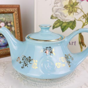 Peut inclure: Une théière en céramique bleu clair avec un couvercle et une anse. La théière est ornée de motifs floraux et de bordures dorées. Une peinture encadrée et une impression florale sont en arrière-plan.