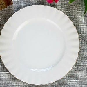 May include: A white ceramic dinner plate with a scalloped edge and a gold rim. The plate is on a gray textured surface.