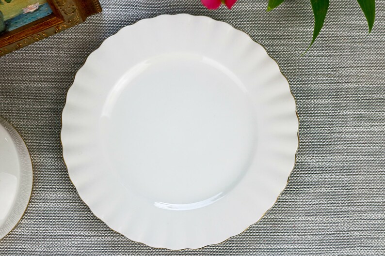 May include: A white ceramic dinner plate with a scalloped edge and a gold rim. The plate is on a gray textured surface.