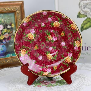 May include: A red and gold floral teacup saucer with a delicate floral pattern. The saucer is on a wooden stand.