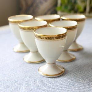 Peut inclure: Ensemble de six coquetiers en porcelaine blanche avec des bordures dorées. Chaque coquetier présente une bande dorée avec un motif de clé grecque. Ils sont disposés sur une surface bleu clair.