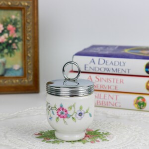 K&ouml;nnte beinhalten: Eine wei&szlig;e Keramik-Eierbecher mit Blumenmuster und einem silbernen Deckel mit einem Ringgriff. Der Eierbecher steht auf einer wei&szlig;en Spitzentischdecke.
