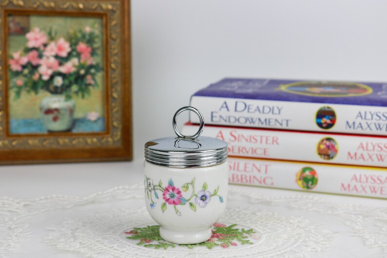 K&ouml;nnte beinhalten: Eine wei&szlig;e Keramik-Eierbecher mit Blumenmuster und einem silbernen Deckel mit einem Ringgriff. Der Eierbecher steht auf einer wei&szlig;en Spitzentischdecke.