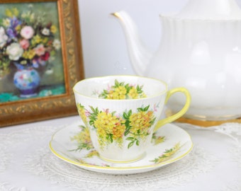 May include: A white teacup and saucer with a yellow floral pattern. The teacup has a delicate handle and a wide rim. The saucer has a matching floral pattern and a yellow rim.