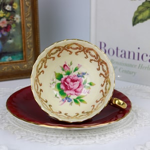 May include: A vintage teacup and saucer set featuring a floral design. The teacup is white with gold trim and a pink rose with green leaves. The saucer is a deep red colour.