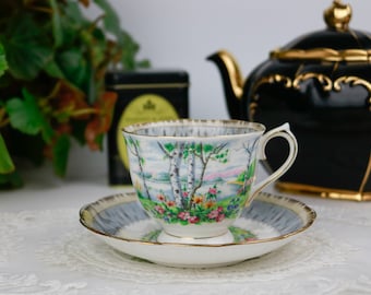 Può includere: Servizio da tè vintage composto da tazza e piattino decorati con un paesaggio con betulle e fiori. La tazza e il piattino hanno bordi dorati. Una teiera nera con dettagli dorati è visibile sullo sfondo.