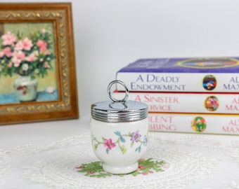 May include: White ceramic egg cup with a floral design and a silver lid with a ring handle. The egg cup is sitting on a white lace doily.