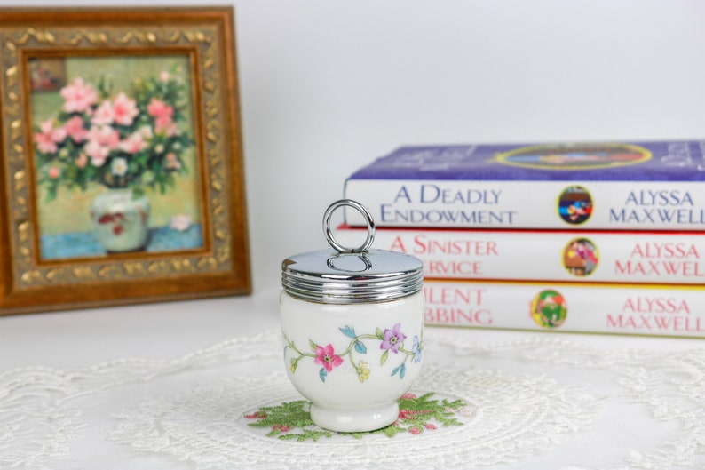 K&ouml;nnte beinhalten: Ein wei&szlig;er Eierbecher aus Keramik mit Blumenmuster und einem silbernen Deckel mit Ringgriff. Der Eierbecher steht auf einer wei&szlig;en Spitzenunterlage.