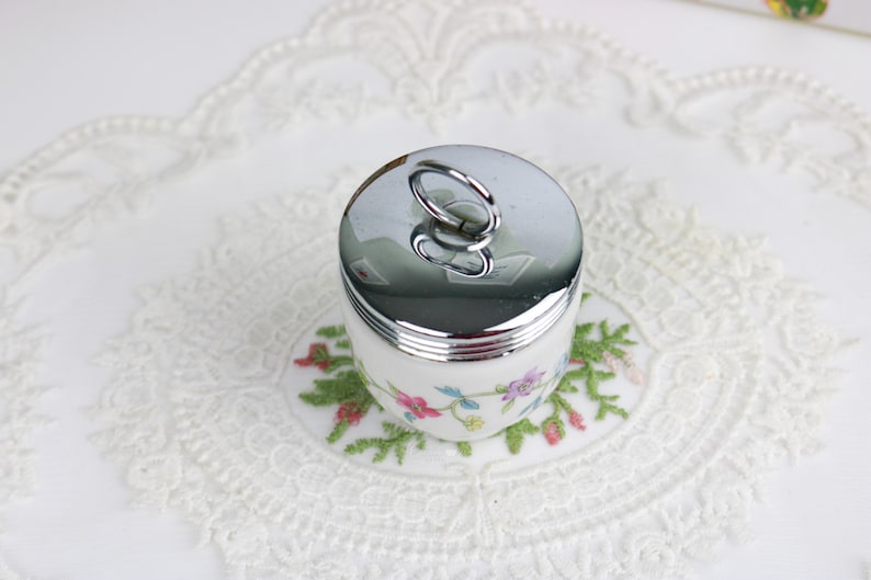 K&ouml;nnte beinhalten: Eine wei&szlig;e Keramik-Eierbecher mit Blumenmuster und einem silbernen Deckel mit einem Schleifenhenkel.