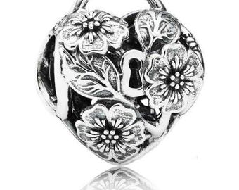 Puede incluir: Un charm de plata en forma de corazón con un diseño floral en relieve que incluye tres flores y hojas. El centro de la pieza presenta un recorte vertical en forma de cerradura y un pequeño aro en la parte superior para sujetarlo a una cadena.