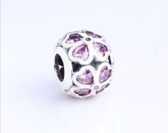 Puede incluir: Un abalorio de metal plateado con un patrón floral repetitivo. Cada forma de flor está incrustada con piedras facetadas de color rosa claro que captan la luz. El abalorio tiene un orificio central para enhebrar en una pulsera o cadena.