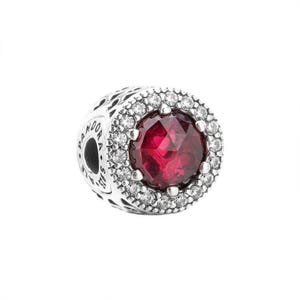 May include: A round silver charm bead featuring a large, faceted, deep red centre stone surrounded by a halo of small, clear, sparkling stones. The side of the silver bead is engraved with the text PANDORA and S925, indicating the metal quality.