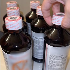 May include: Several dark brown bottles with white labels and red and white security seals. The bottles are being held by a hand. The labels have text that is difficult to read. The bottles appear to contain a liquid.