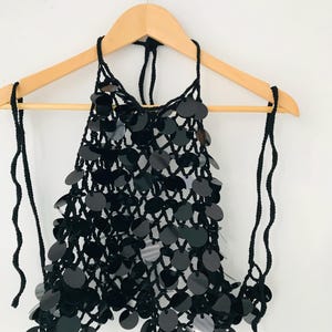 May include: Black halter top with large, round sequins. The top is made of a crocheted net and hangs on a wooden hanger. The halter straps are made of black yarn. The sequins are shiny and reflect light.