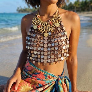 May include: A halter top constructed from brown cord featuring numerous circular metallic silver discs and a prominent gold-toned statement necklace with coral-like branches and coin charms, paired with a floral sarong in shades of orange and blue.