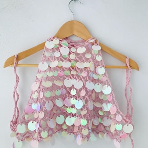 May include: A light pink crochet halter top featuring a loose, open-knit mesh construction adorned with numerous circular, iridescent sequins that reflect light with a pearlescent sheen, suspended from a wooden hanger by thin crocheted ties.