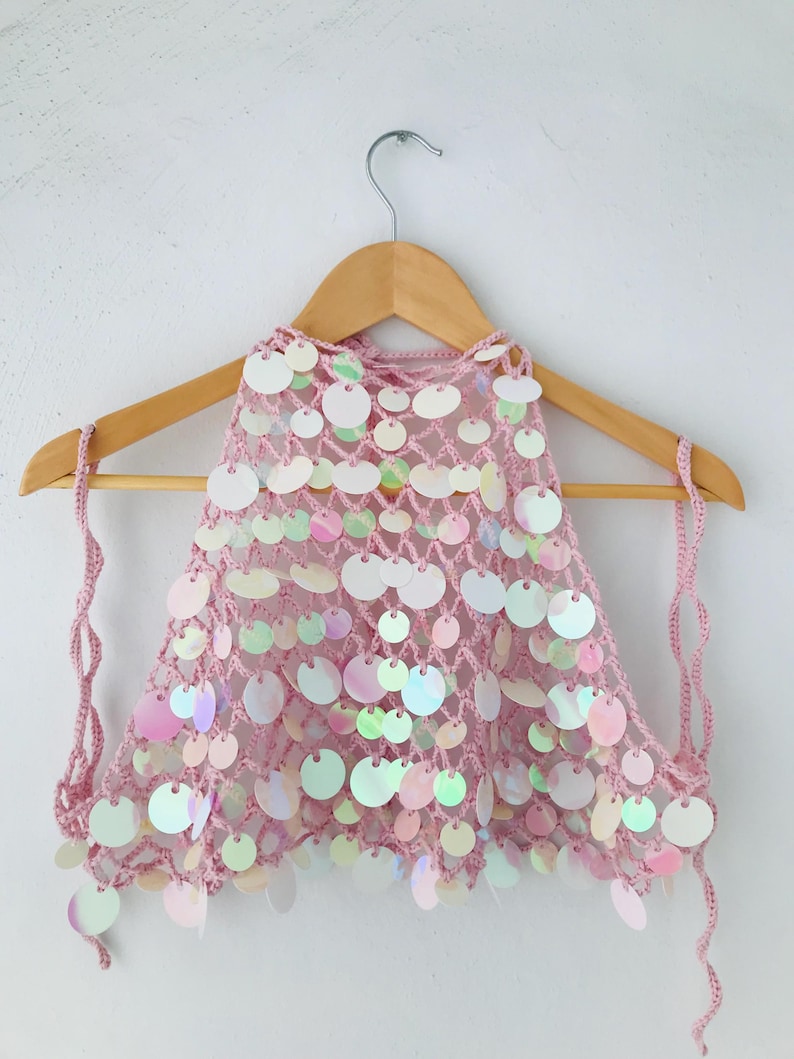 May include: A light pink crochet halter top featuring a loose, open-knit mesh construction adorned with numerous circular, iridescent sequins that reflect light with a pearlescent sheen, suspended from a wooden hanger by thin crocheted ties.