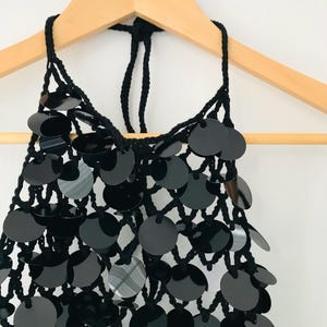 May include: A black halter top with a net-like design, embellished with many round, shiny black sequins. The top is hanging from a wooden hanger, highlighting its intricate details and texture. The garment's design is suitable for special occasions.