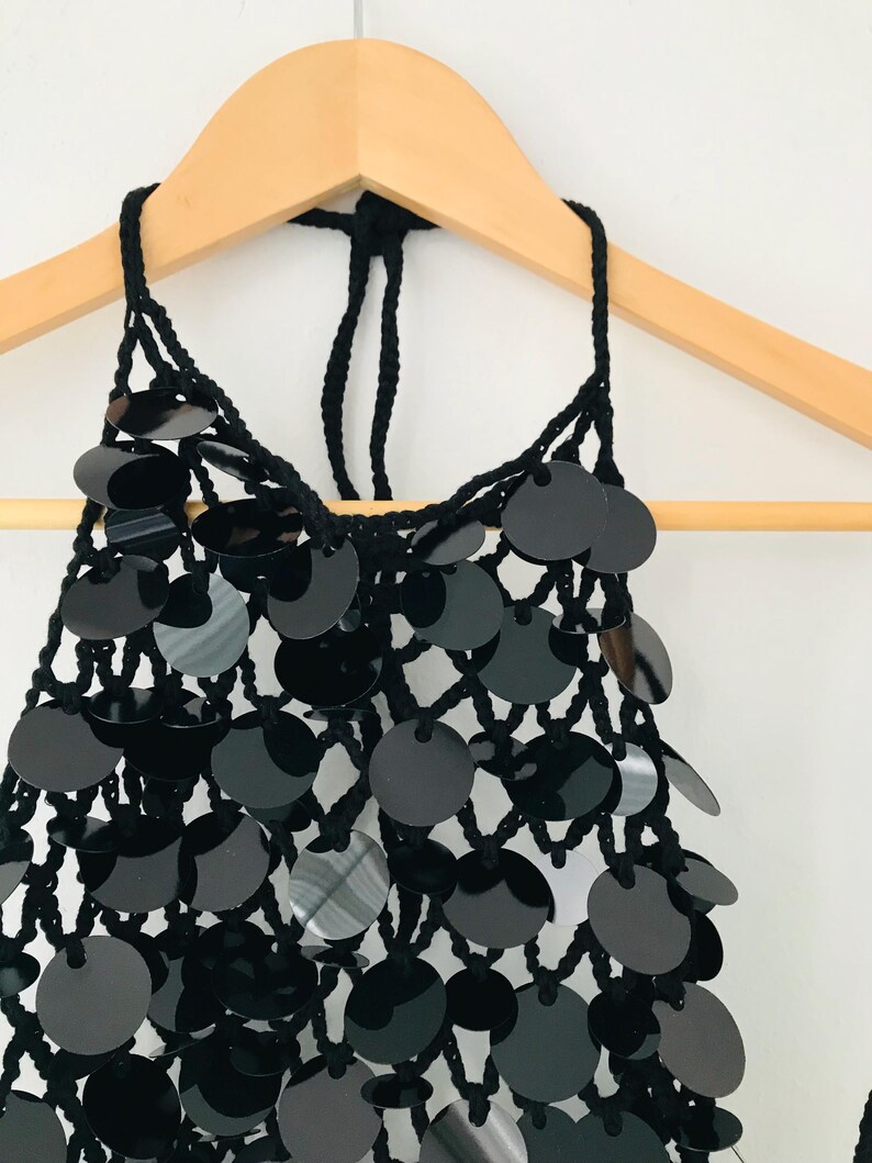 May include: A black halter top with a net-like design, embellished with many round, shiny black sequins. The top is hanging from a wooden hanger, highlighting its intricate details and texture. The garment's design is suitable for special occasions.