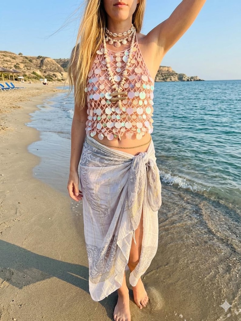 May include: A sleeveless halter top made of light pink crochet mesh adorned with iridescent circular sequins. The top is paired with a light gray sarong featuring a subtle white and gold floral pattern, tied at the waist with a side knot.