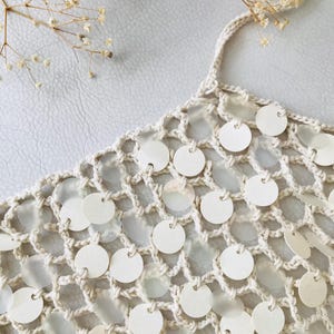 May include: A cream-colored crochet top featuring an open-knit mesh pattern. The garment is embellished with numerous small, circular, semi-translucent white discs attached to the yarn intersections throughout the entire piece.