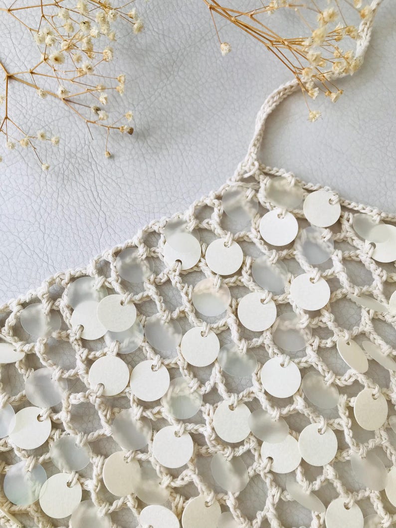 May include: A cream-colored crochet top featuring an open-knit mesh pattern. The garment is embellished with numerous small, circular, semi-translucent white discs attached to the yarn intersections throughout the entire piece.