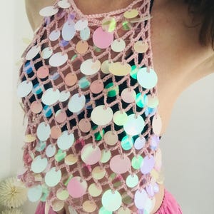 May include: A light pink crochet halter top featuring a wide-mesh net design adorned with numerous circular, iridescent sequins that reflect various colors. The garment is worn over a black base layer and paired with a bright pink pleated skirt.