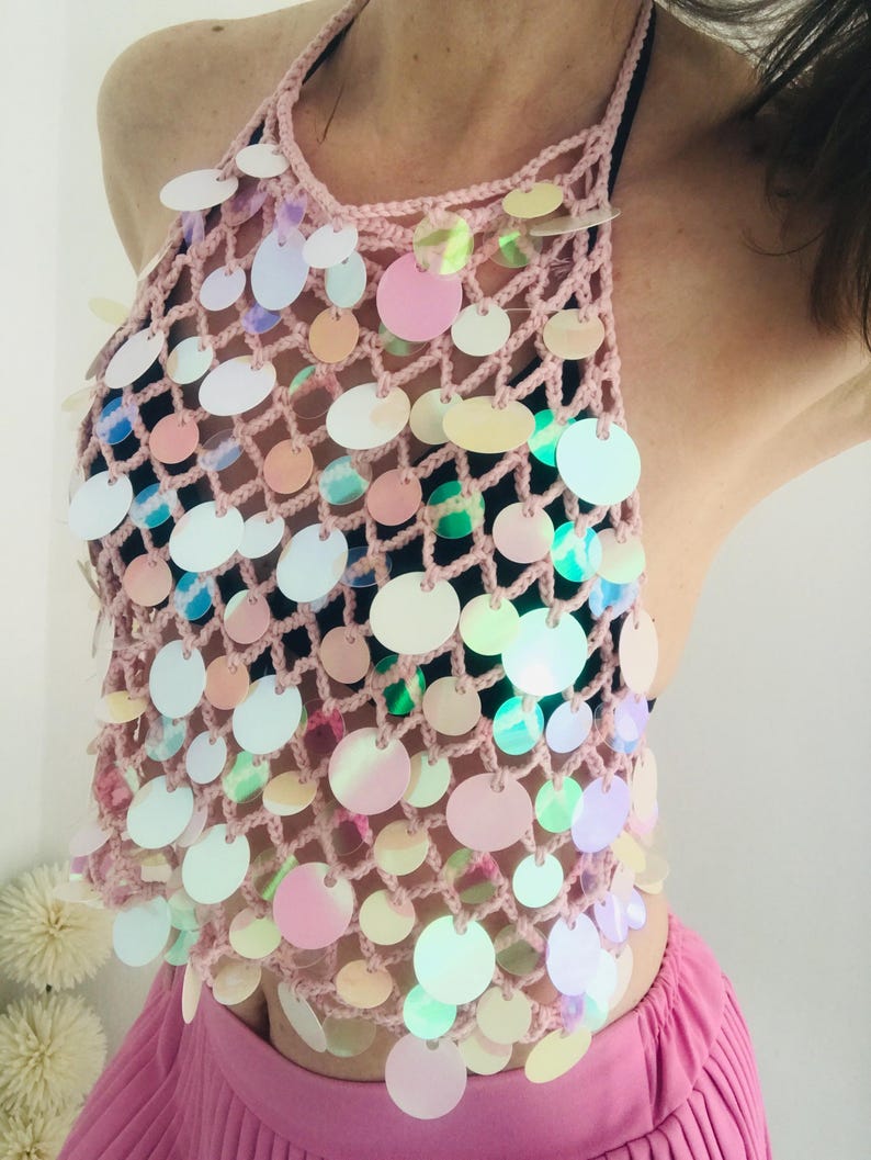 May include: A light pink crochet halter top featuring a wide-mesh net design adorned with numerous circular, iridescent sequins that reflect various colors. The garment is worn over a black base layer and paired with a bright pink pleated skirt.