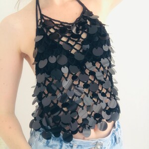 May include: Black halter top with a crochet design and large, round, reflective sequins. The top features a halter neck and a series of sequins cascading down the front. The top is paired with light blue denim jeans.
